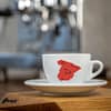 Eine weiße Cappuccino-Tasse bedruckt mit der roten Silhouette von Spanien. In schwarz darauf ein Stier und Symbole für Berge und Wind.