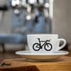 Eine weiße Cappuccino-Tasse bedruckt mit einem Triathlon-Rad auf der Vorderseite auf einer hölzernen Oberfläche. Im Hintergrund eine glänzende Siebträger-Kaffeemaschine.