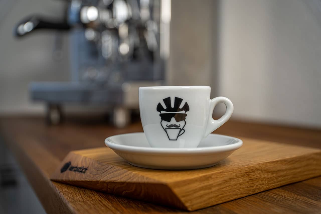 Eine Espresso-Tasse bedruckt mit dem Gesicht eines Fahrradfahrers mit Helm, Brille und Schnauzbart.