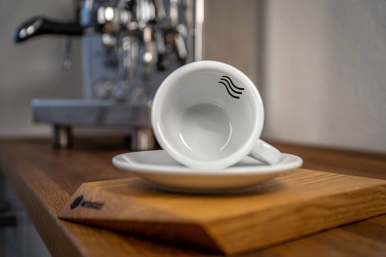 Eine weiße Cappuccino-Tasse bedruckt mit einem Wellen-Symbol auf der Innenseite, im Hintergrund eine glänzende Siebträger-Kaffeemaschine.