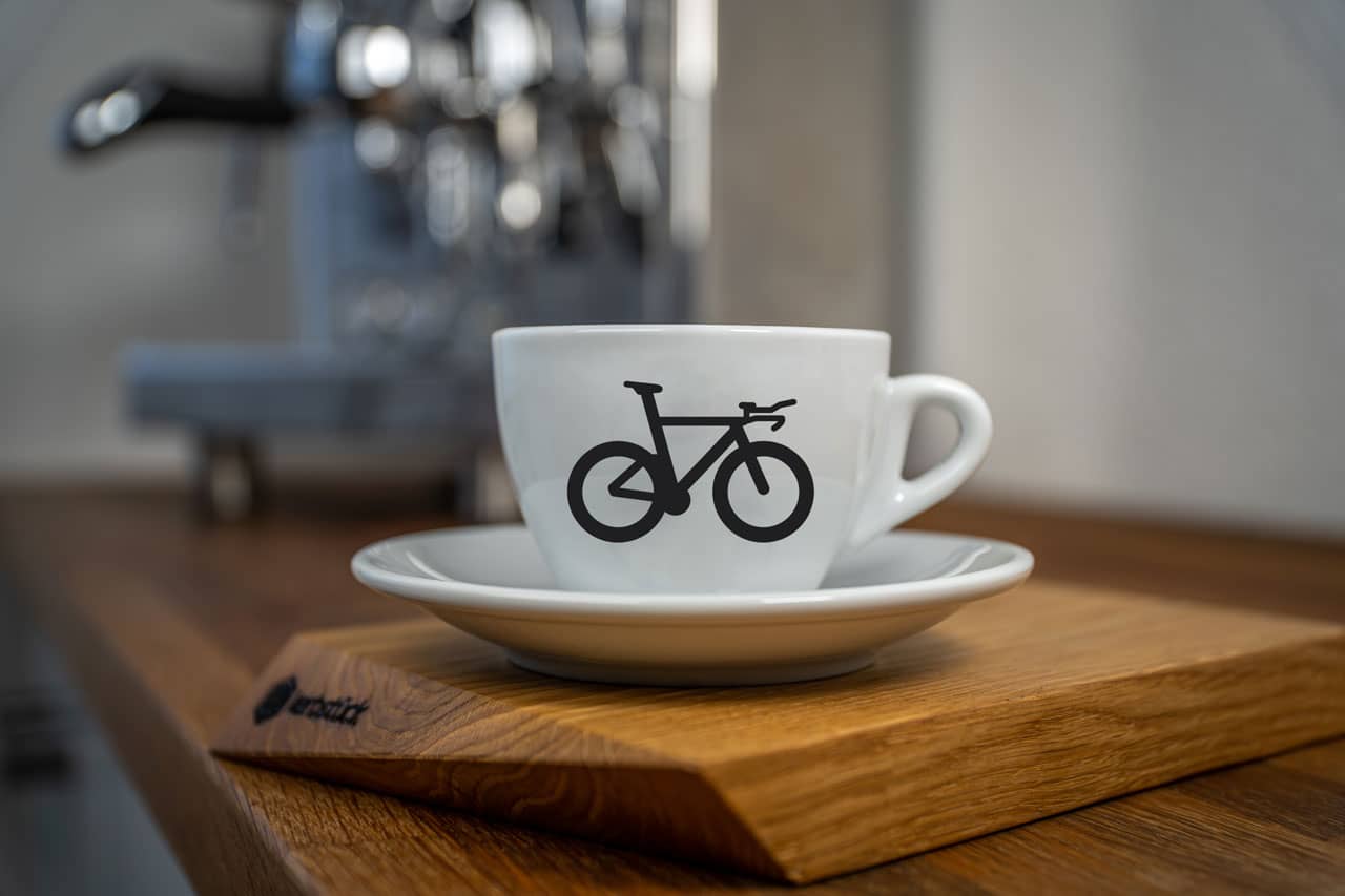 Eine weiße Cappuccino-Tasse bedruckt mit einem Triathlon-Rad auf der Vorderseite auf einem Holzbrett, im Hintergrund eine glänzende Siebträger-Kaffeemaschine.