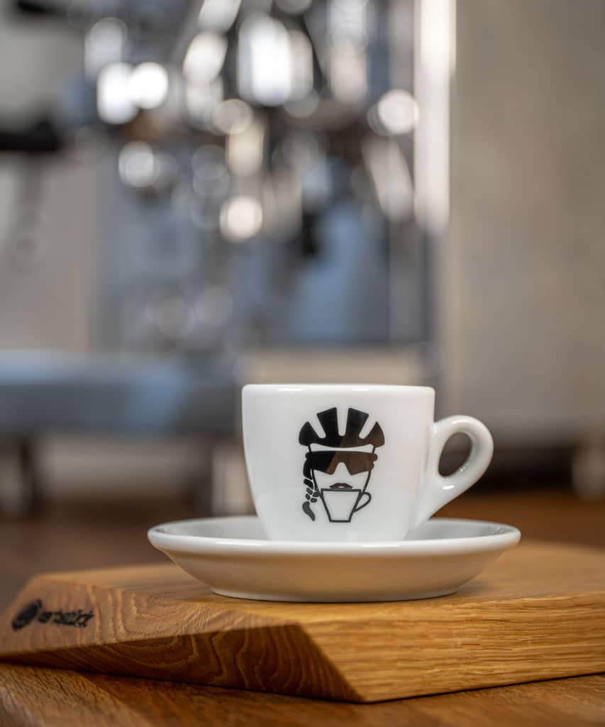 Eine weiße Espresso-Tasse bedruckt mit einem stilisierten Kopf einer Radfahrerin mit Helm, Sonnenbrille und Zopf, die aus einer Kaffeetasse trinkt.