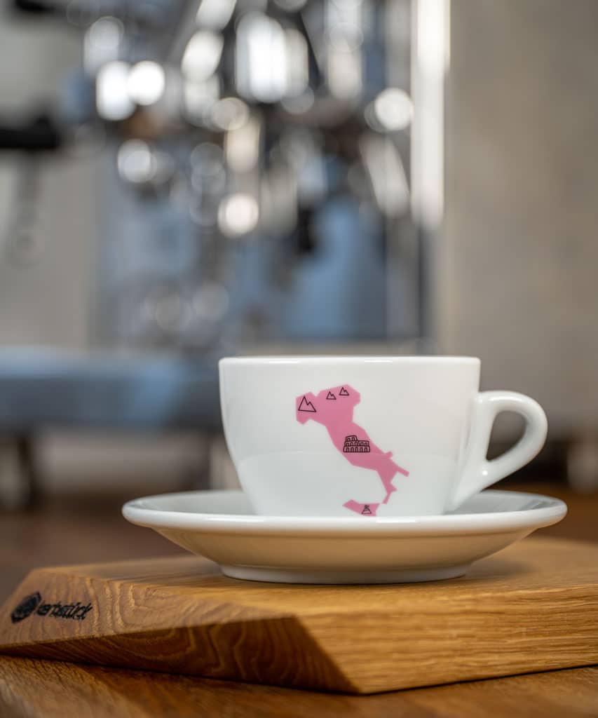 Eine weiße Cappuccino-Tasse bedruckt mit der rosanen Silhouette von Italien. In schwarz darauf das Kolosseum und Symbole für Berge und Wind.