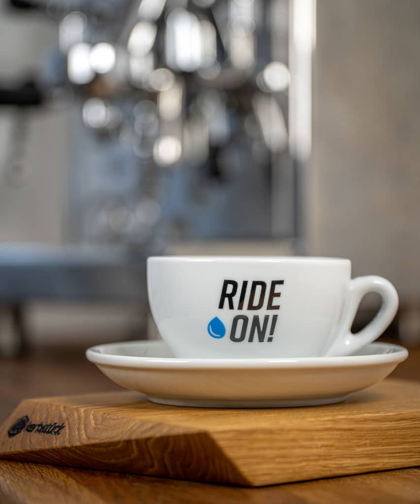 Eine weiße Cappuccino-Tasse mit dem Schriftzug Ride on und einem blauen Wassertropfen