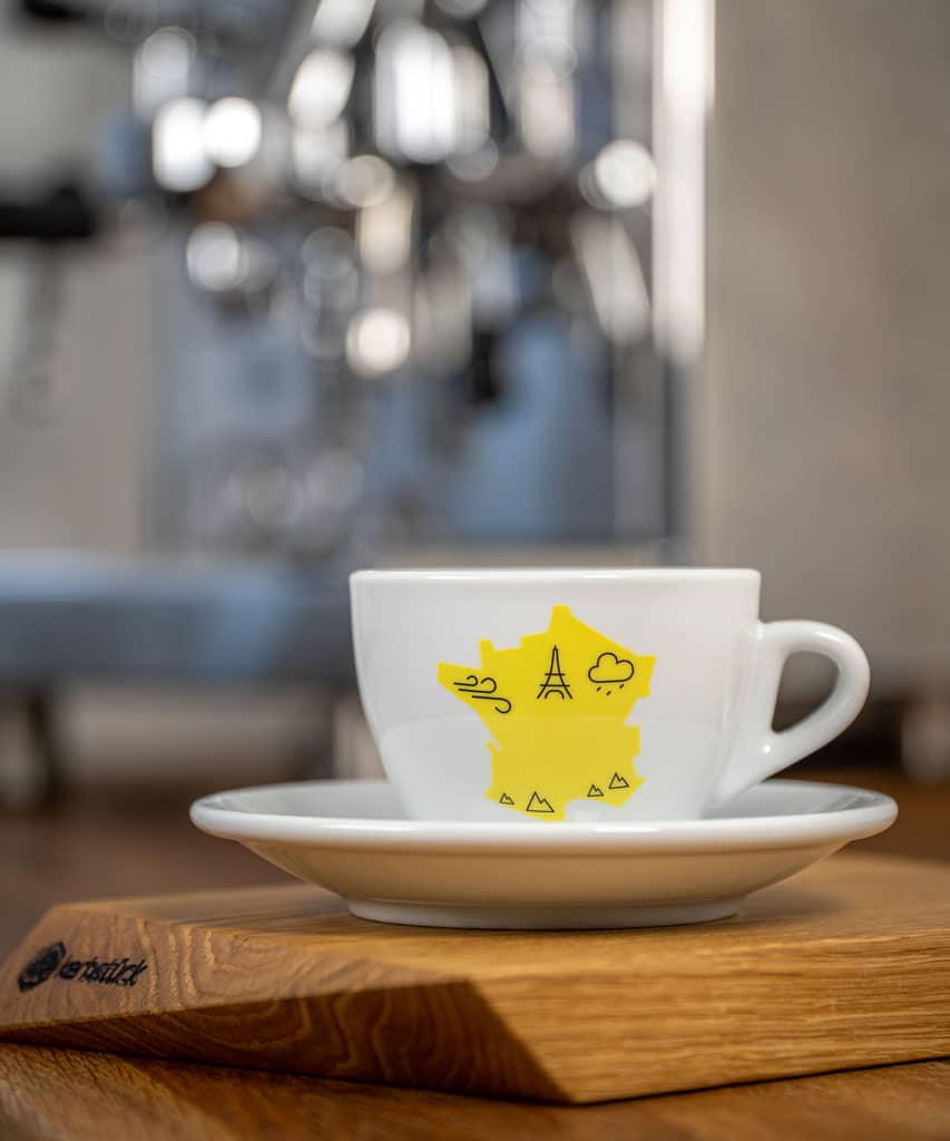 Eine weiße Cappuccino-Tasse bedruckt mit der gelben Silhouette von Frankreich. In schwarz darauf der Eifelturm und Symbole für Berge, Wind und Regen.
