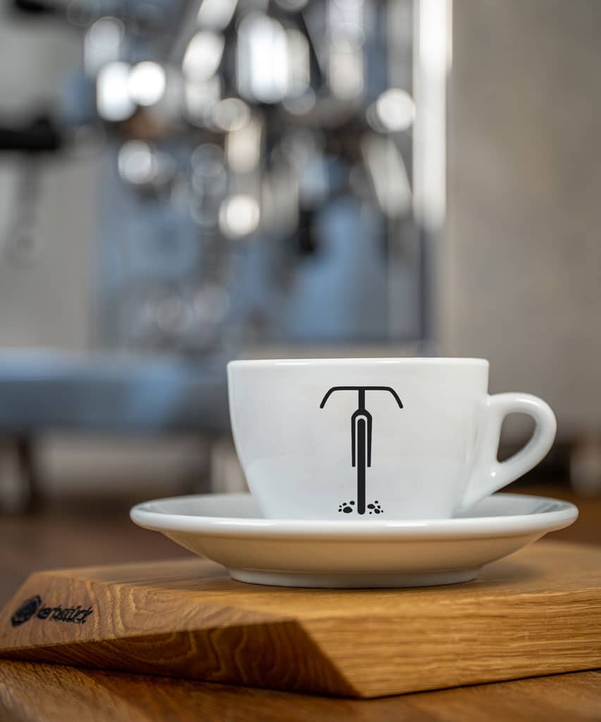Eine weiße Cappuccino-Tasse bedruckt mit der schwarzen Silhouette eines Gravel-Rades. Im Hintergrund eine glänzende Siebträger-Kaffeemaschine.