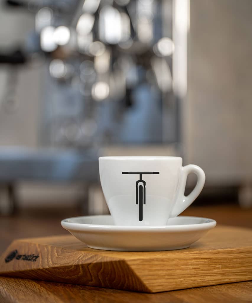 Eine weiße Espresso-Tasse bedruckt mit der schwarzen Moutainbike-Silhouette. Im Hintergrund eine glänzende Siebträger-Kaffeemaschine.