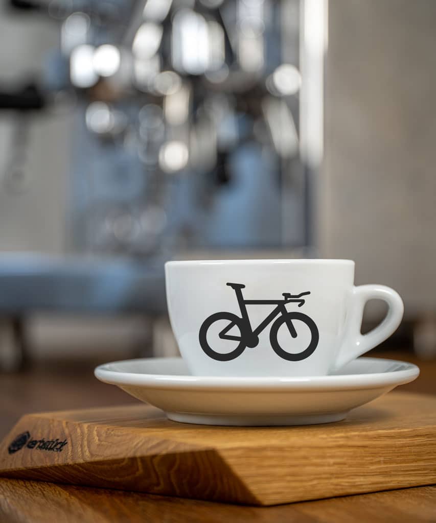 Eine weiße Cappuccino-Tasse bedruckt mit einem Triathlon-Rad auf der Vorderseite auf einer hölzernen Oberfläche. Im Hintergrund eine glänzende Siebträger-Kaffeemaschine.