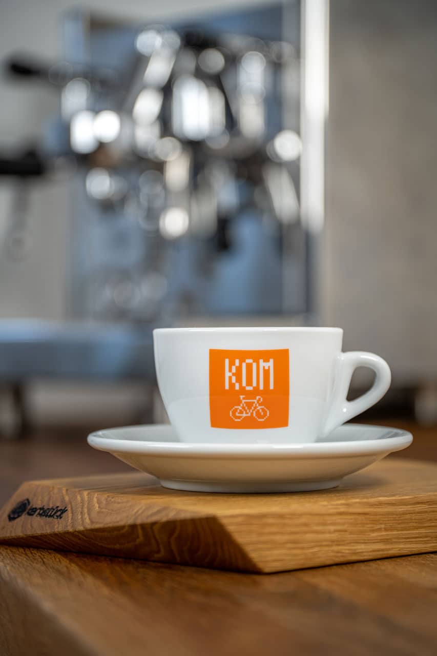 Eine weiße Cappuccino-Tasse bedruckt mit einem orangenen Block mit weißer Aufschrift KOM und einem verpixelten Rennrad.