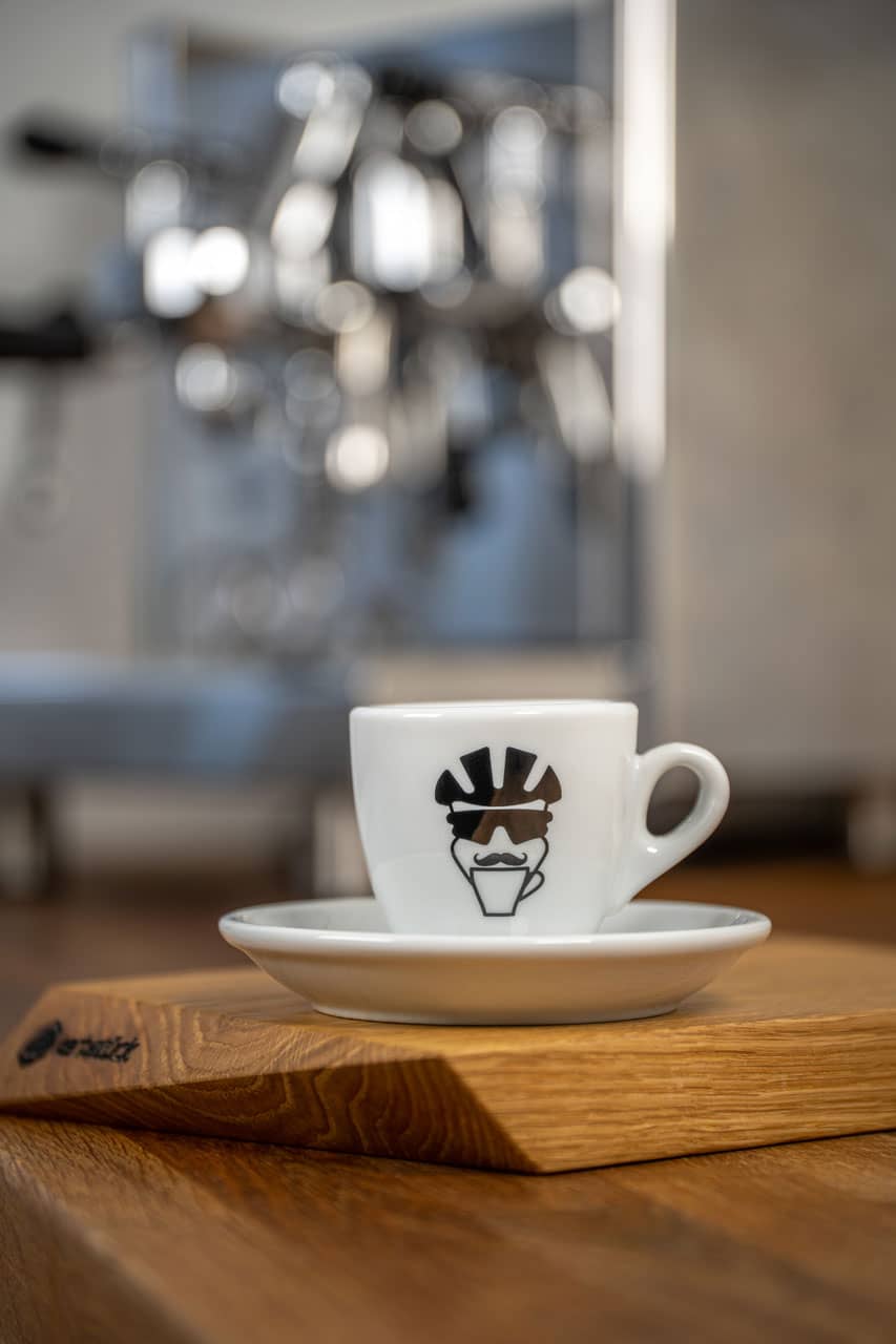Eine Espresso-Tasse bedruckt mit dem Gesicht eines Fahrradfahrers mit Helm, Brille und Schnauzbart.