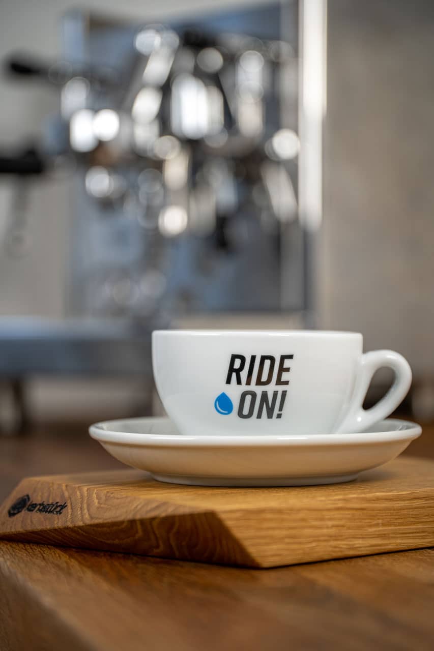 Eine weiße Cappuccino-Tasse mit dem Schriftzug Ride on und einem blauen Wassertropfen