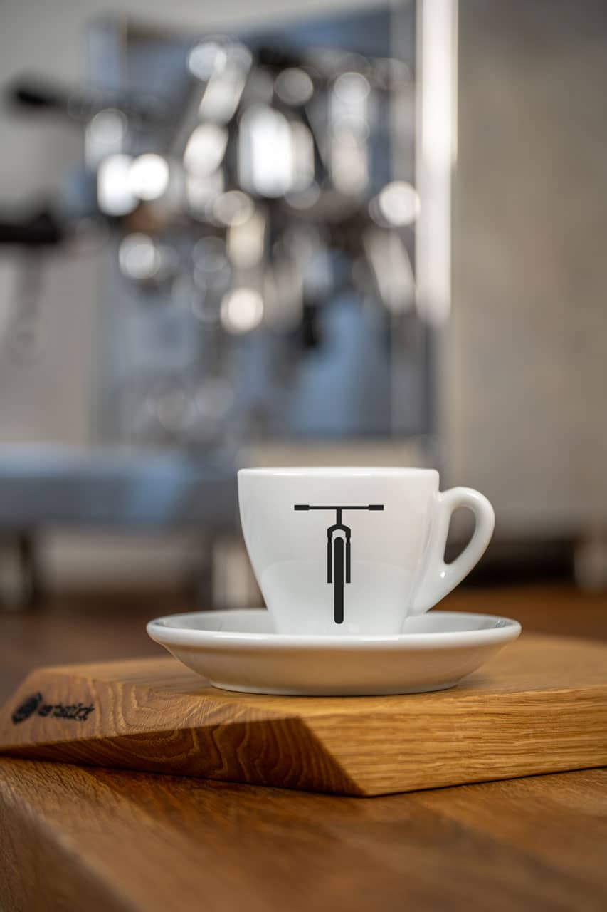 Eine weiße Espresso-Tasse bedruckt mit der schwarzen Moutainbike-Silhouette. Im Hintergrund eine glänzende Siebträger-Kaffeemaschine.