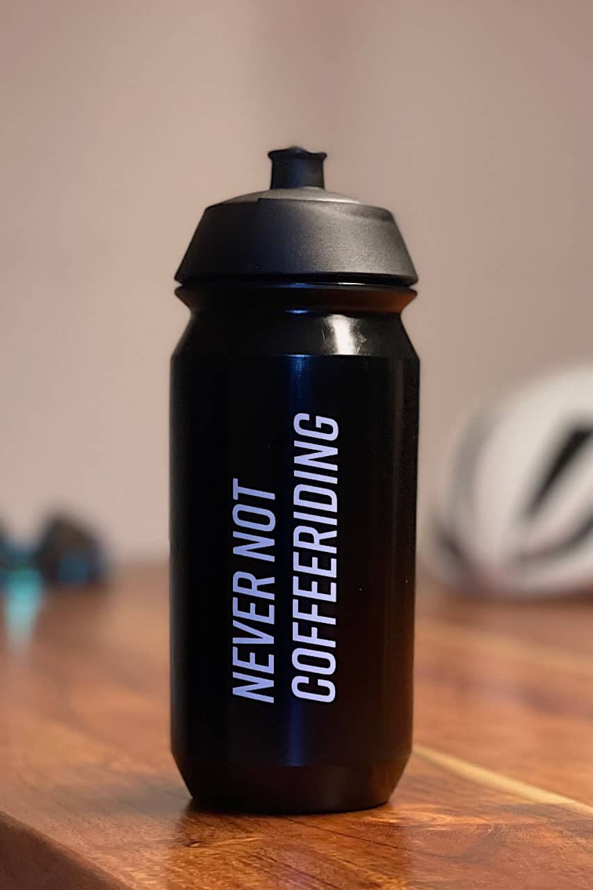 Eine schwarze Fahrrad-Trinkflasche mit dem Spruch "Never not Coffeeriding" auf einem Tisch vor einem verschwommenen Hintergrund.