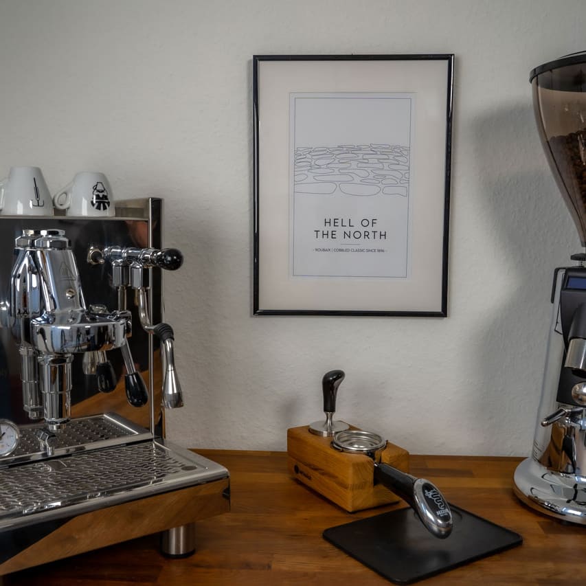 Das Poster "Hell of the North" hängt im Bilderrahmen an der Wand zwischen einer Kaffeemaschine und einer Kaffeemühle.
