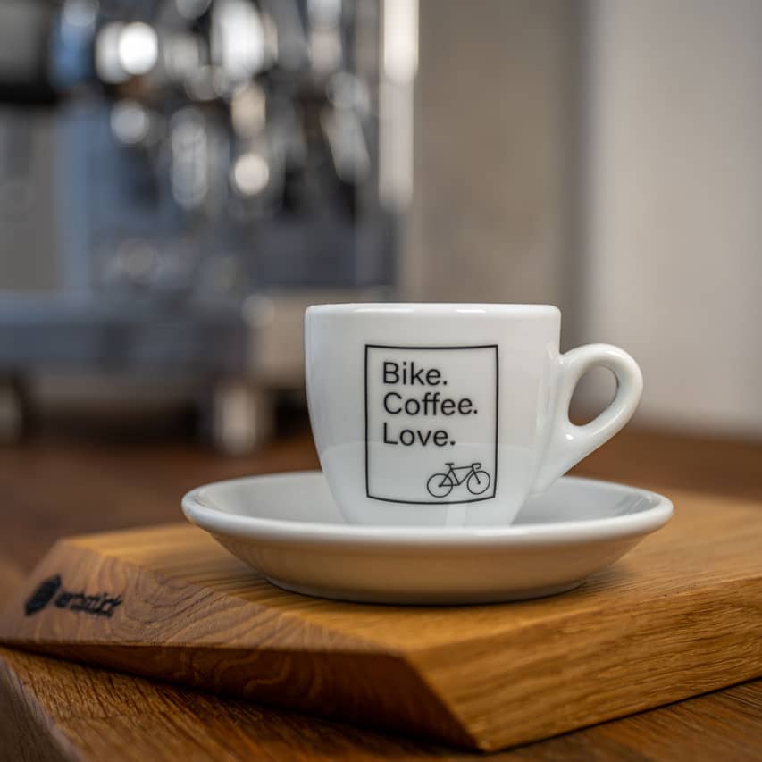 Eine weiße Espresso-Tasse bedruckt mit dem Slogan "Bike Coffee Love" und einem stilisierten Rennrad in einem schwarzen Kasten.