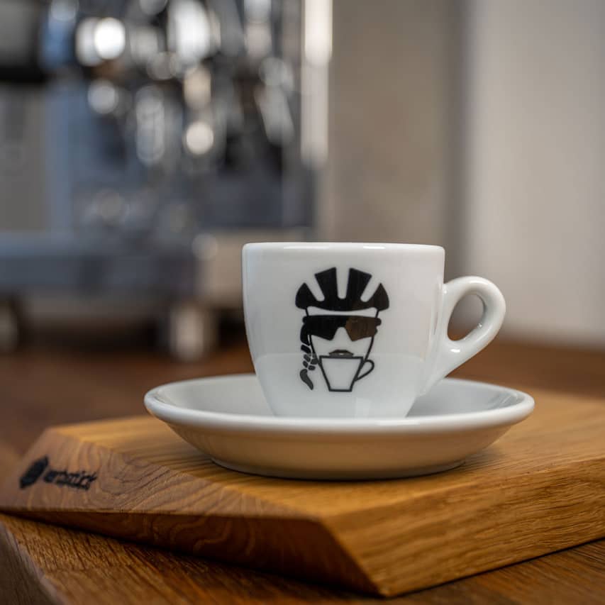 Eine weiße Espresso-Tasse bedruckt mit einem stilisierten Kopf einer Radfahrerin mit Helm, Sonnenbrille und Zopf, die aus einer Kaffeetasse trinkt.