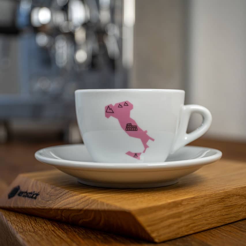 Eine weiße Cappuccino-Tasse bedruckt mit der rosanen Silhouette von Italien. In schwarz darauf das Kolosseum und Symbole für Berge und Wind.