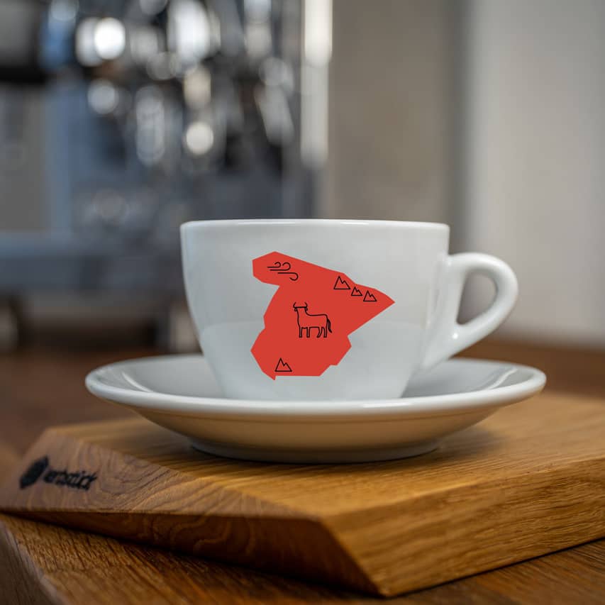 Eine weiße Cappuccino-Tasse bedruckt mit der roten Silhouette von Spanien. In schwarz darauf ein Stier und Symbole für Berge und Wind.