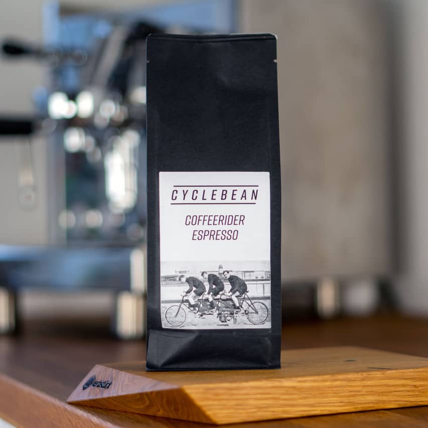 Eine Espresso-Packung steht auf einer Arbeitsfläche vor einer Kaffeemaschine. Auf den Label sind drei Radfahrer zu sehen.